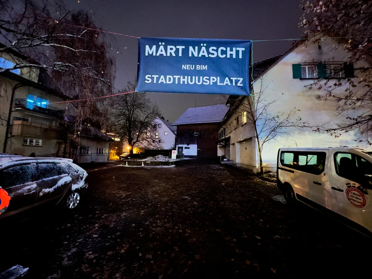 Man sieht ein Banner, das über einem Platz aufgespannt ist. Darauf steht: «Märt Näscht neu bim Stadthuusplatz».